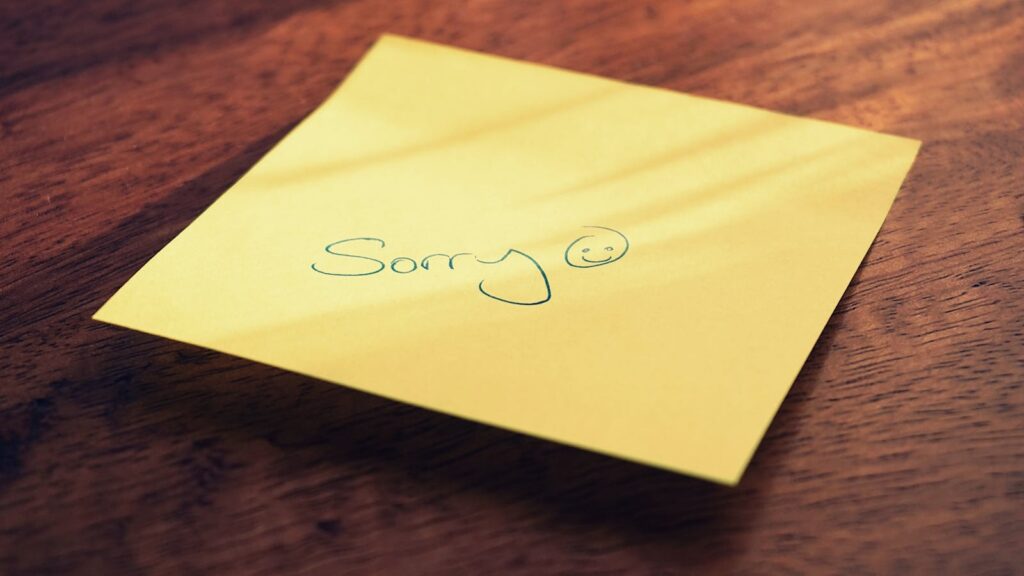 A yellow sticky note with a handwritten apology message and smiley face on a wooden surface.